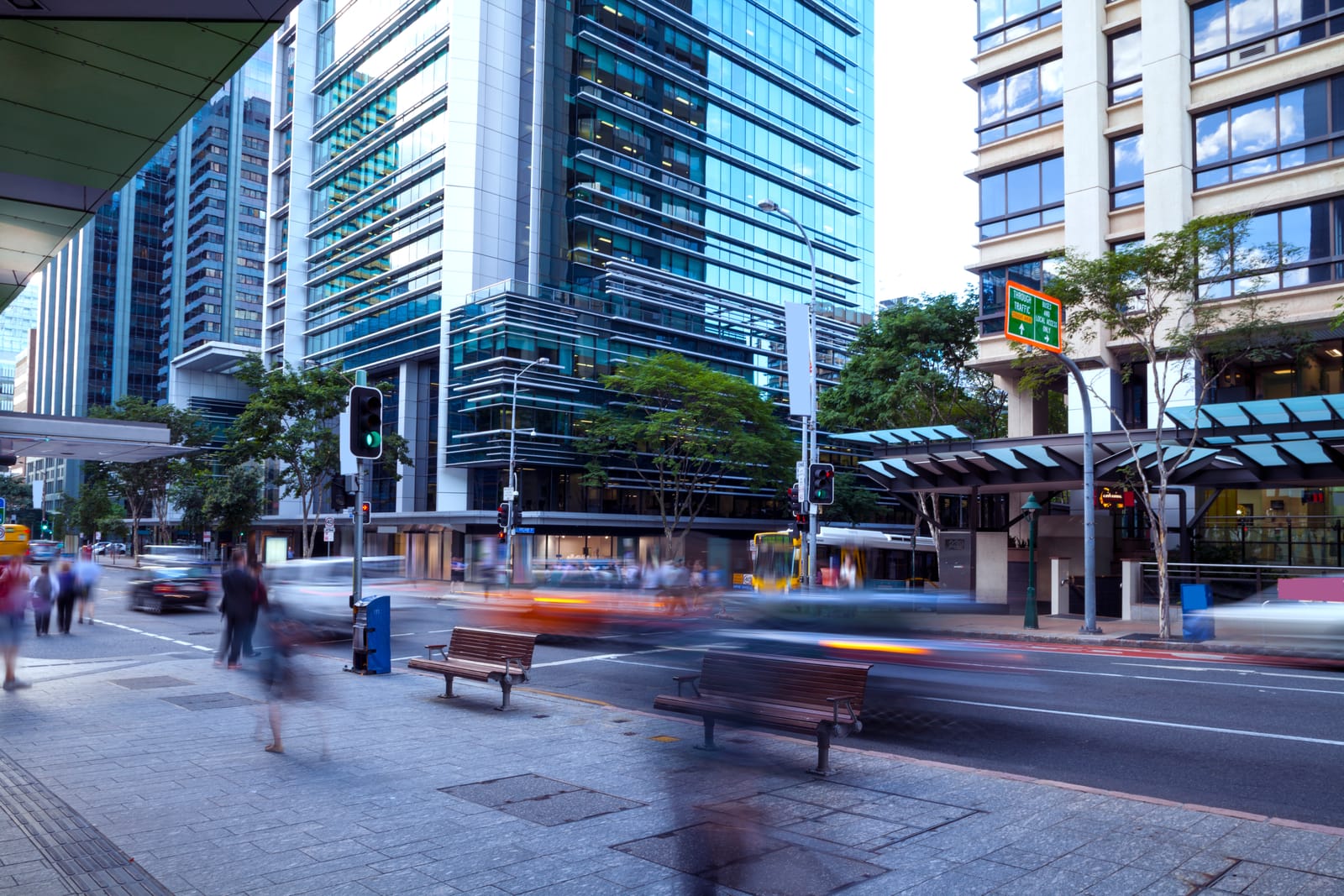Pedestrians and traffic blur past glass office towers and street trees on a Brisbane CBD street